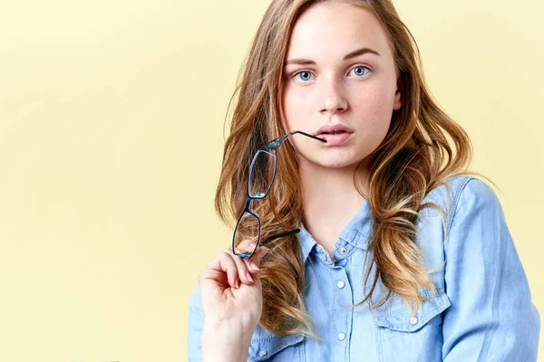 Hermosa adolescente con cabello de jengibre, pecas y ojos azules sosteniendo gafas de lectura, mujer joven con gafas sobre fondo amarillo —  Fotos de Stock