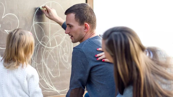 Padres jóvenes con su linda niña dibujando en pizarra. Familia divirtiéndose al aire libre, sosteniendo tiza y dibujo . —  Fotos de Stock
