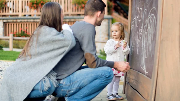 Giovani genitori con la loro carina bambina che disegna sulla lavagna. Famiglia che si diverte all'aperto, tenendo gesso e disegno . — Foto Stock