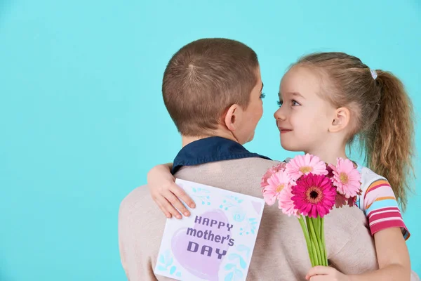 Happy Mother's Day. Cute little girl giving mom mothers day card and bouquet of pink gerbera daisies. Mother and daughter concept on candy blue pastel background.