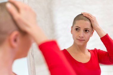 Young adult female cancer patient looking in the mirror, stroking her new short hair.  clipart