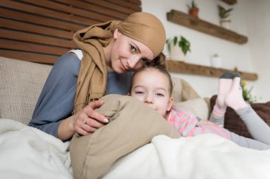 Young adult female cancer patient spending time with her daughter at home, relaxing on the couch. Cancer and family support concept. clipart