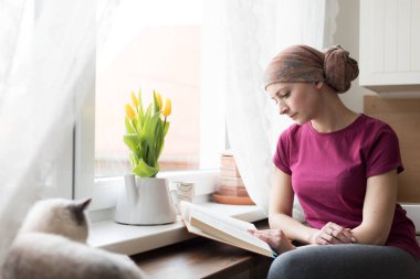 Young adult female cancer patient wearing headscarf sitting in the kitchen with her pet cat, relaxing by reading a book. clipart