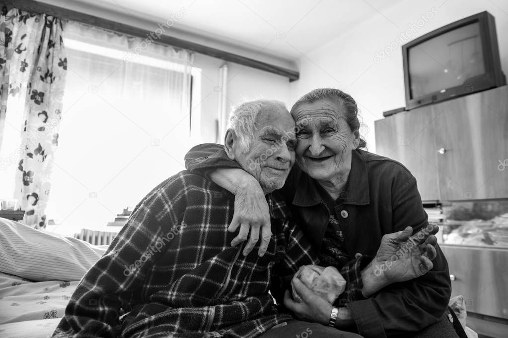 Cute 80 plus year old senior married couple hugging and smiling portrait. Black and white waist up image of happy elderly couple sitting on a bed in a nursing home.