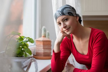 Young positive adult female cancer patient sitting in the kitchen by a window, smiling and looking at the camera. clipart