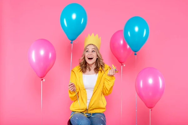 Funny girl in birthday hat, balloons and blowout horn on pastel pink background. Attractive trendy teenager celebrating birthday.