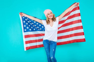 Amerika'yı seviyorum. Mutlu genç gülümseyen kadın kot pantolon ve Amerikan bayrağı tutarak ve kamera pastel mavi arka plan karşı dururken bakarak beyaz Tshirt.