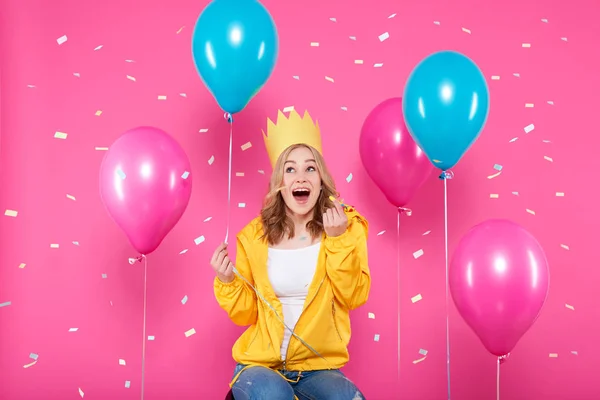 Funny girl in birthday hat, balloons and flying confetti on pastel pink background. Attractive trendy teenager celebrating birthday.