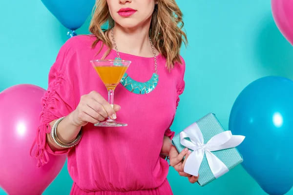 Gorgeous trendy young woman in party outfit holding cocktail and a present. Birthday Party concept on pastel blue background.