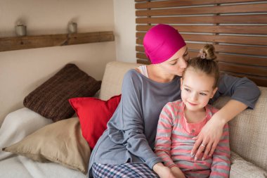 Young adult female cancer patient spending time with her daughter at home, relaxing on the couch. Cancer and family support concept. clipart