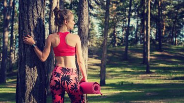 Genç atletik kadın akıllı saati ve yoga mat tutan bir ormandaki yerleştirmek. Zihin ve beden kavramı kopya alanı ile. Yoga egzersiz.