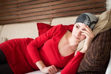 Young adult female cancer patient relaxing on a couch, watching television. Tired, exhausted, depressed cancer patient. clipart