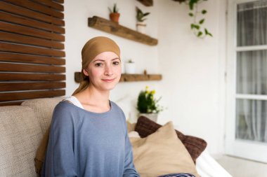 Oturma odasında, gülümseyen ve kameraya bakarak genç olumlu yetişkin kadın kanser hasta oturma portre.