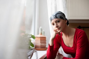 Young adult female cancer patient wearing headscarf sitting by the window in the kitchen looking at camera. clipart