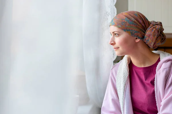 年轻成年女性癌症患者戴头巾和浴衣坐在厨房里看窗外 — 图库照片