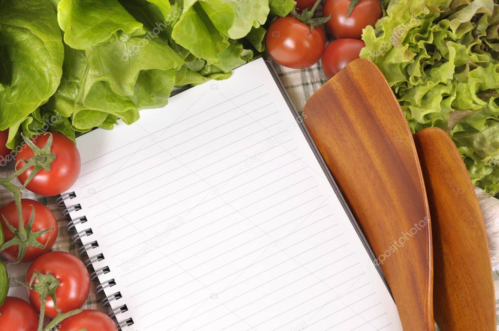 Selection of salad vegetables with blank recipe book or shopping list and wooden serving spoons.