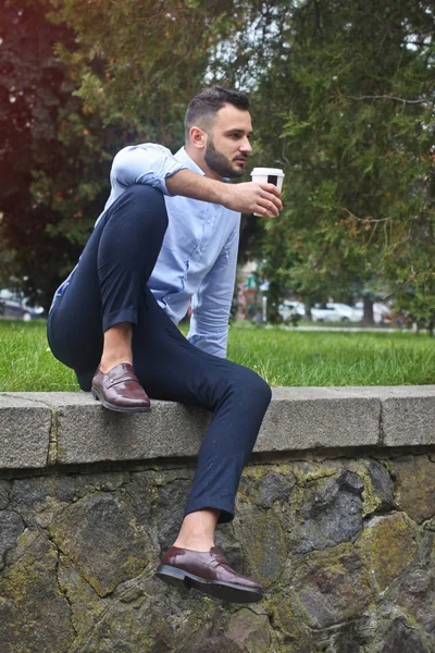 Retrato de un tipo con una camisa sentado en un parque en una ciudad europea. Un hombre en el fondo de árboles y edificios. Fotos de stock —  Fotos de Stock