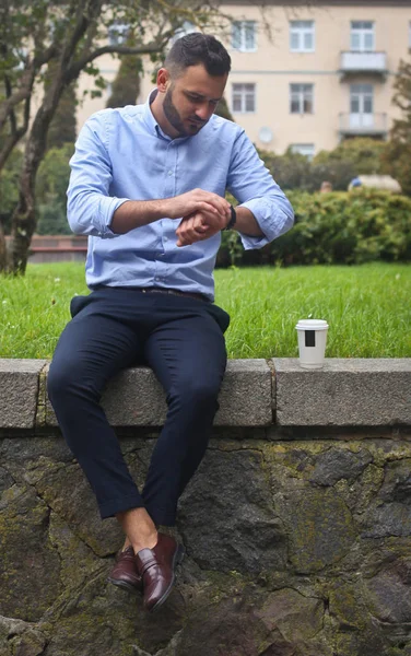 Retrato de un tipo con una camisa sentado en un parque en una ciudad europea. Un hombre mirando el reloj contra el fondo de árboles y edificios. Fotos de stock —  Fotos de Stock