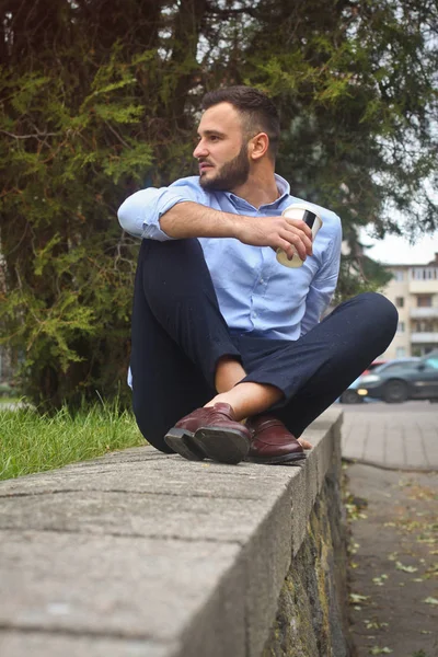 Retrato de un tipo con una camisa sentado en un parque en una ciudad europea. Un hombre en el fondo de árboles y edificios. Fotos de stock —  Fotos de Stock