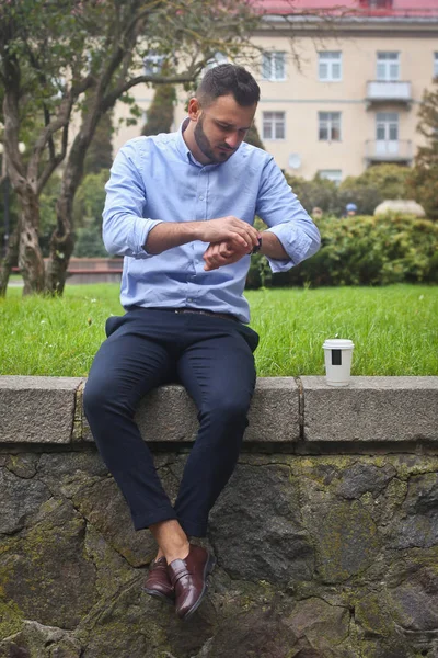 Retrato de un tipo con una camisa sentado en un parque en una ciudad europea. Un hombre mirando el reloj contra el fondo de árboles y edificios. Fotos de stock —  Fotos de Stock