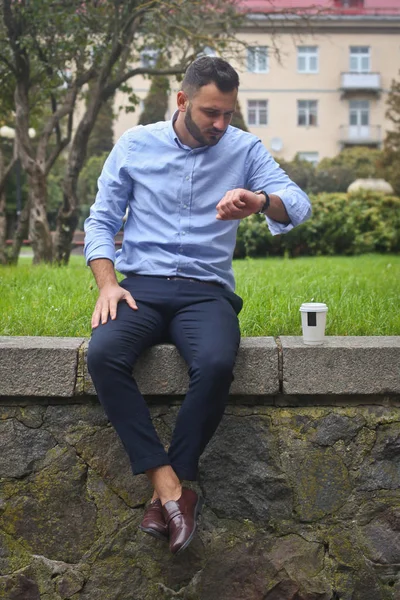 Retrato de un tipo con una camisa sentado en un parque en una ciudad europea. Un hombre mirando el reloj contra el fondo de árboles y edificios. Fotos de stock —  Fotos de Stock