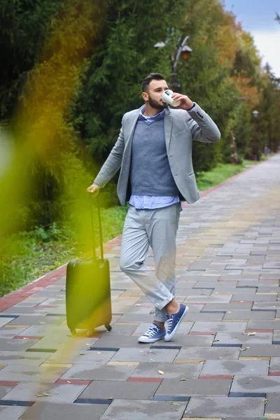 Tipo vestido a la moda está viajando con el equipaje en el fondo de una carretera y árboles. El hombre se apresura en su vuelo y vierte café, té. Foto de stock para el diseño —  Fotos de Stock