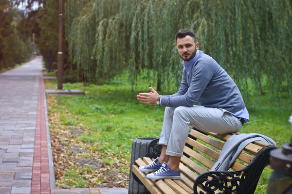 Un joven sexy sentado en un banco en un parque otoñal de verano. Un hombre contemplando nuevas ideas mientras camina. Campo europeo. Foto de stock —  Fotos de Stock