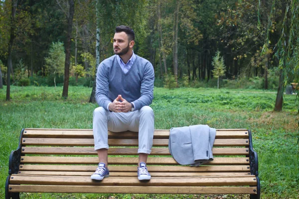 Un joven sexy sentado en un banco en un parque otoñal de verano. Un hombre contemplando nuevas ideas mientras camina. Campo europeo. Foto de stock —  Fotos de Stock