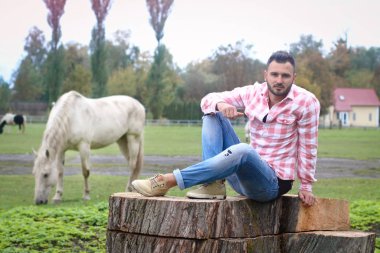 Genç yakışıklı Kovboy. Çiftçi çiftliğinde büyük bir kütükte oturuyor. Kırsal alanlar, kırsal alanlar. Ağaçlar, tarlalar, çiftlik. Desenleme için stok fotoğrafları