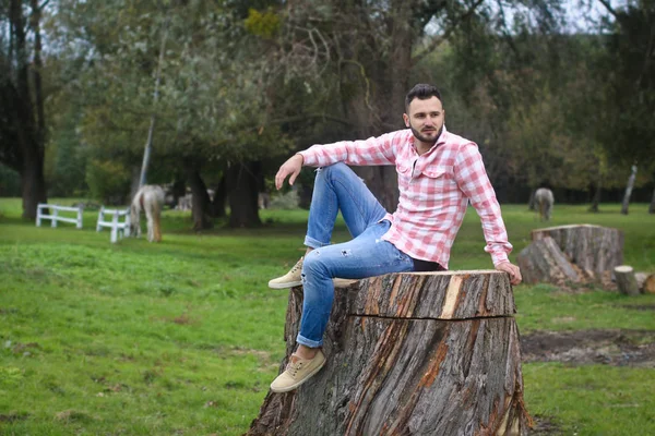 Joven chico apuesto Vaquero. granjero está sentado en un gran tocón en su rancho. Paisajes rurales, campo. Árboles, campo, granja. Fotos de stock para desig — Foto de Stock