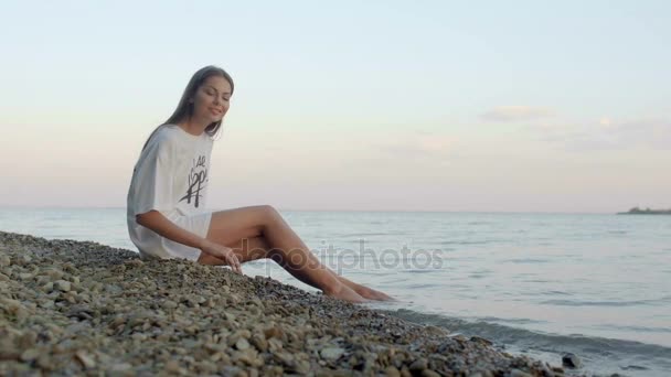 Femme réfléchie assise sur la plage de galets et touchant des rochers — Video