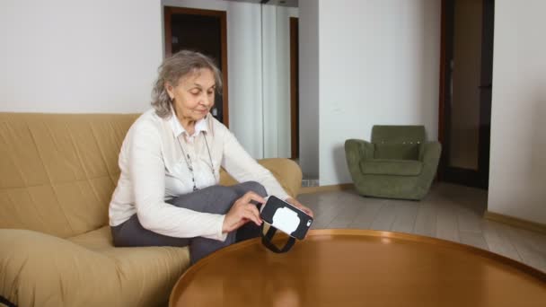 Femme âgée est assis sur le canapé et mettre des lunettes VR avec téléphone portable. Belle femme âgée en blanc utilisant des lunettes VR 360 à la maison . — Video