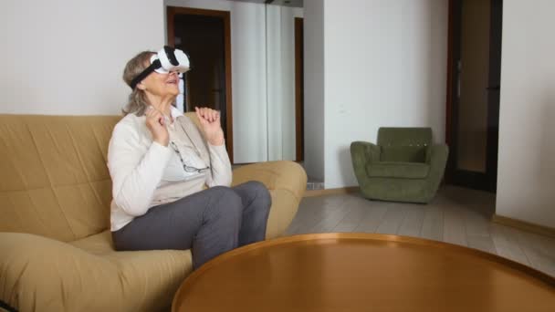 Sorrindo, a mulher sênior está sentada no sofá em óculos VR com celular. Mulher Sênior de boa aparência em branco usando óculos VR 360 em casa . — Vídeo de Stock
