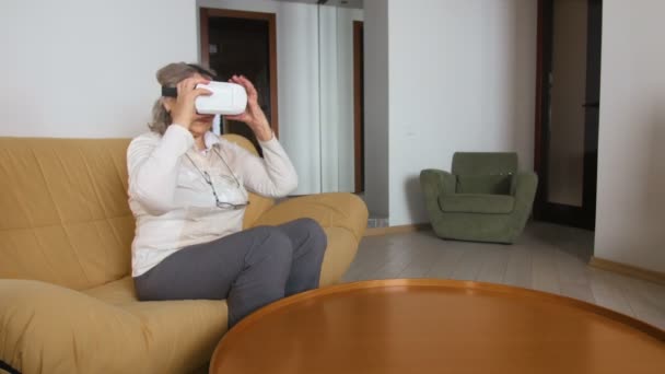 Seniorin sitzt auf der Couch und spielt Vr Brille. gut aussehende Seniorin in Weiß mit 360-Grad-Brille zu Hause. — Stockvideo