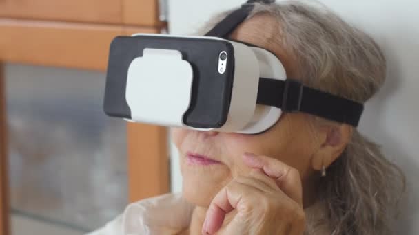 Gros plan de la femme âgée est assis sur le canapé et de mettre des lunettes VR avec téléphone portable. Belle femme âgée en blanc utilisant des lunettes VR 360 à la maison . — Video