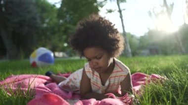 Afro-Amerikalı kızın yakın çekimleri. Parkta, yatak örtüsünün üzerinde uzanıyor ve telefonda çizgi film izliyor.
