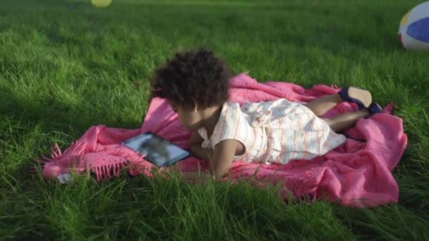 Küçük Afro-Amerikalı kız, parka uzanmış, yatak örtüsüne uzanmış, tablet üzerinde çizgi film izliyor. — Stok video
