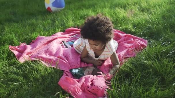 Little african-american girl is watching cartoons on tablet in park — Stock Video