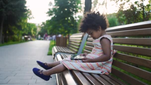 Niña afroamericana está sentada en el banco en el parque y viendo dibujos animados en la tableta — Vídeos de Stock