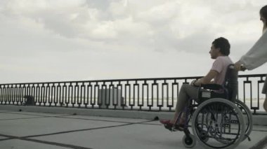 Genç engelli adam tekerlekli sandalyeyle yürüyor, kadın onu yuvarlıyor. Birlikte nehir kenarındaki parkta dinleniyorlar.