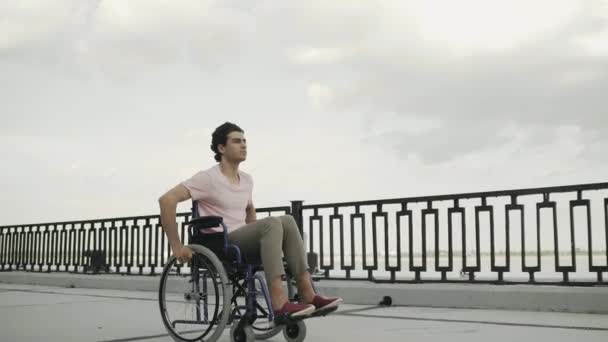 L'uomo invalido seduto sulla sedia a rotelle cavalca lungo l'argine — Video Stock