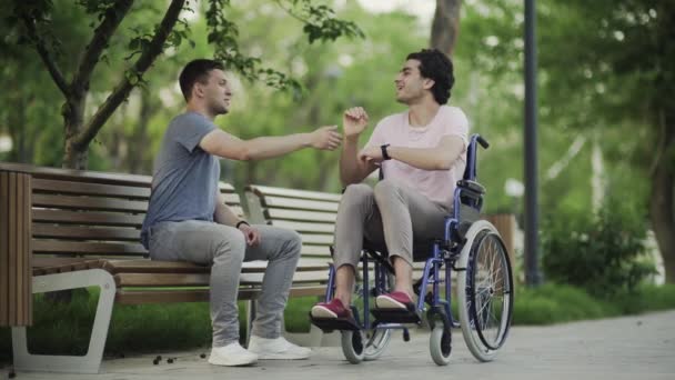 Молодой человек в инвалидной коляске встречает своего друга в парке . — стоковое видео