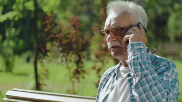 Fešák starší muž mluví po telefonu na lavičce v parku — Stock video