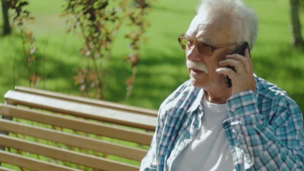 Hombre mayor enojado hablando por teléfono en el banco en el parque — Vídeos de Stock