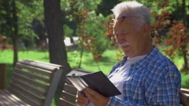 Bıyıklı yaşlı bir adam kitapta komik anlar okuyor. Yaşlı adam parkta bankta oturuyor ve kitap okuyor.