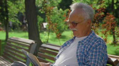 Yaşlı bir adam bankta oturuyor ve parkta komik bir kitap okuyor.