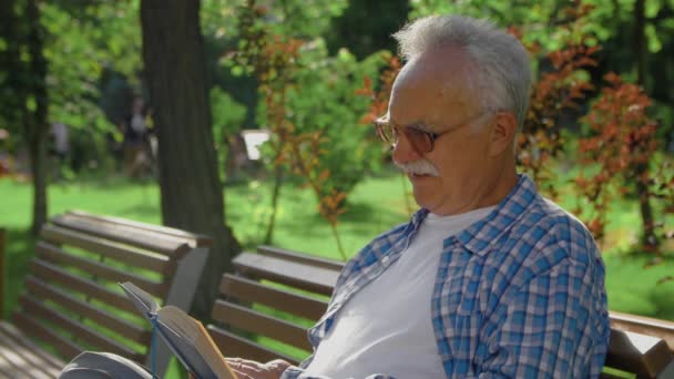 Een oudere man zit op de bank en leest een grappig boek in het park. — Stockvideo