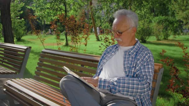 Een oudere man zit op de bank en leest boek in het park — Stockvideo