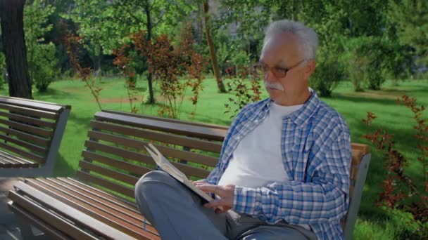 Ένας ηλικιωμένος κάθεται στον πάγκο και διαβάζει ένα αστείο βιβλίο στο πάρκο.. — Αρχείο Βίντεο