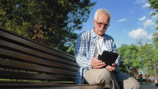 Viejo con gafas y bigote está sentado en el banco en el parque y la lectura de libros electrónicos — Vídeos de Stock